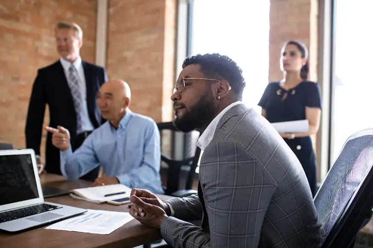 A small team meets around a table in a modern office, discussing work while looking at a laptop.